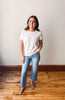 Woman wearing a white t-shirt and blue jeans standing on a wooden floor with a plain wall background