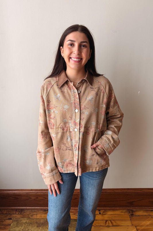 A woman smiling while wearing a beige button-up shirt with a pink and blue floral pattern, paired with blue jeans, standing against a plain beige wall with a wooden baseboard and floor.