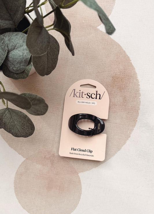 A black flat cloud clip by Kit's on its original beige cardboard packaging, styled on a textured beige surface with a plant in the background.