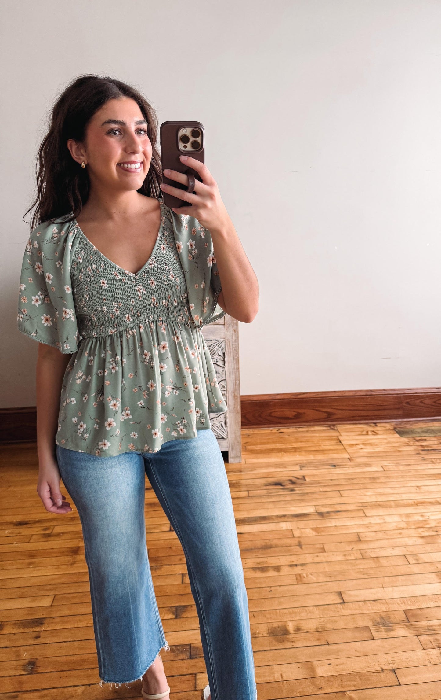 Woman taking a mirror selfie wearing a green floral blouse and blue jeans.
