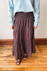 A person wearing a long brown tiered maxi skirt with a small white floral pattern, paired with a light blue sweater and brown sandals, standing on a wooden floor against a plain wall.