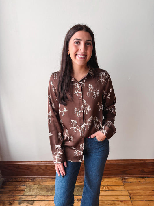 A woman with long dark hair stands indoors wearing a brown button-up shirt with a white horse pattern and blue jeans, posing against a plain off-white wall with a wooden baseboard and wood floor.