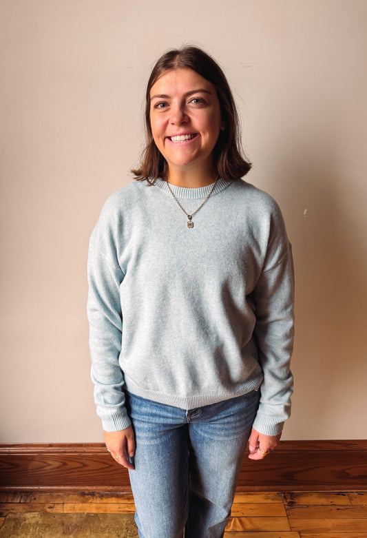 A smiling woman with shoulder-length brown hair stands indoors wearing a light heather gray crewneck sweatshirt and blue jeans against a plain beige wall with a wooden baseboard.