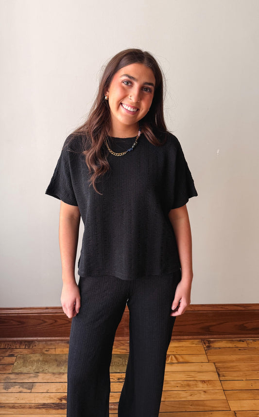 Woman wearing a black t-shirt and black pants standing against a plain wall.