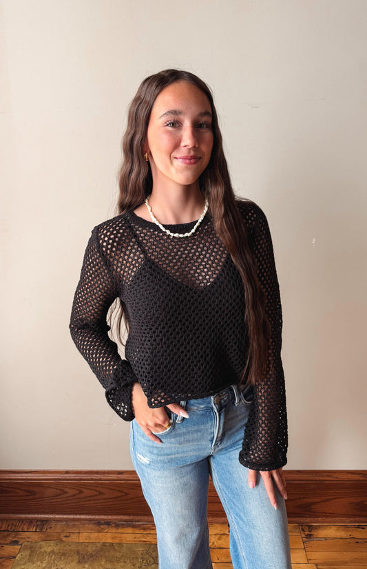 Woman wearing a black mesh top and blue jeans against a plain wall.