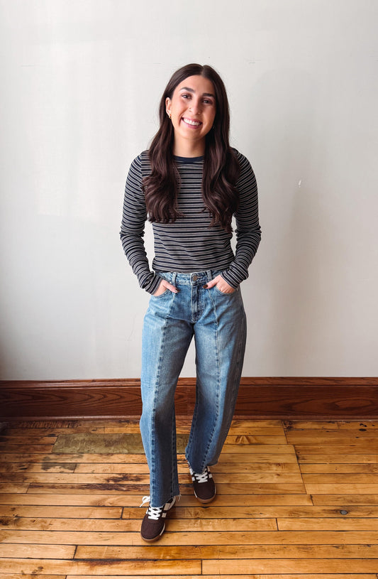 A woman stands on a wooden floor against a plain off-white wall, wearing a black and white striped long-sleeve top, light-wash blue jeans, and dark brown sneakers.