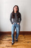 A woman stands on a wooden floor against a plain off-white wall, wearing a black and white striped long-sleeve top, light-wash blue jeans, and dark brown sneakers.