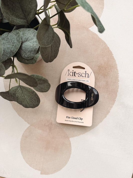 A black flat cloud hair clip attached to a cream-colored Kit-sch retail card, styled on a beige surface with a green plant in the background.