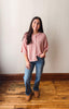 A woman stands in a room with a light beige wall and wooden floor, wearing a pink short-sleeve knit cardigan, blue jeans, and brown boots.