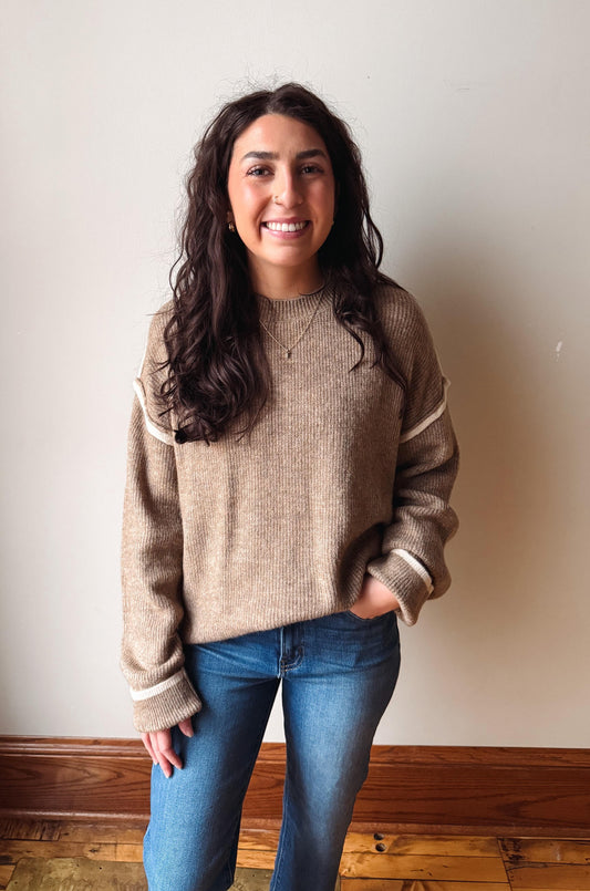 A woman with long dark wavy hair stands indoors wearing a beige knit sweater with cream ribbed trim on the shoulders and cuffs, paired with blue jeans, against a plain off-white wall.