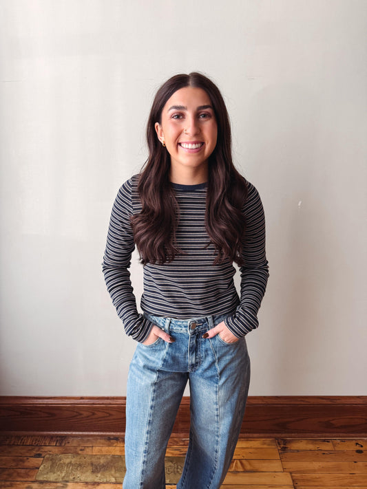 A smiling young woman with long dark brown hair stands indoors wearing a black and gray striped long-sleeve top and light blue jeans, with her hands in her pockets, against a plain off-white wall and wooden floor.