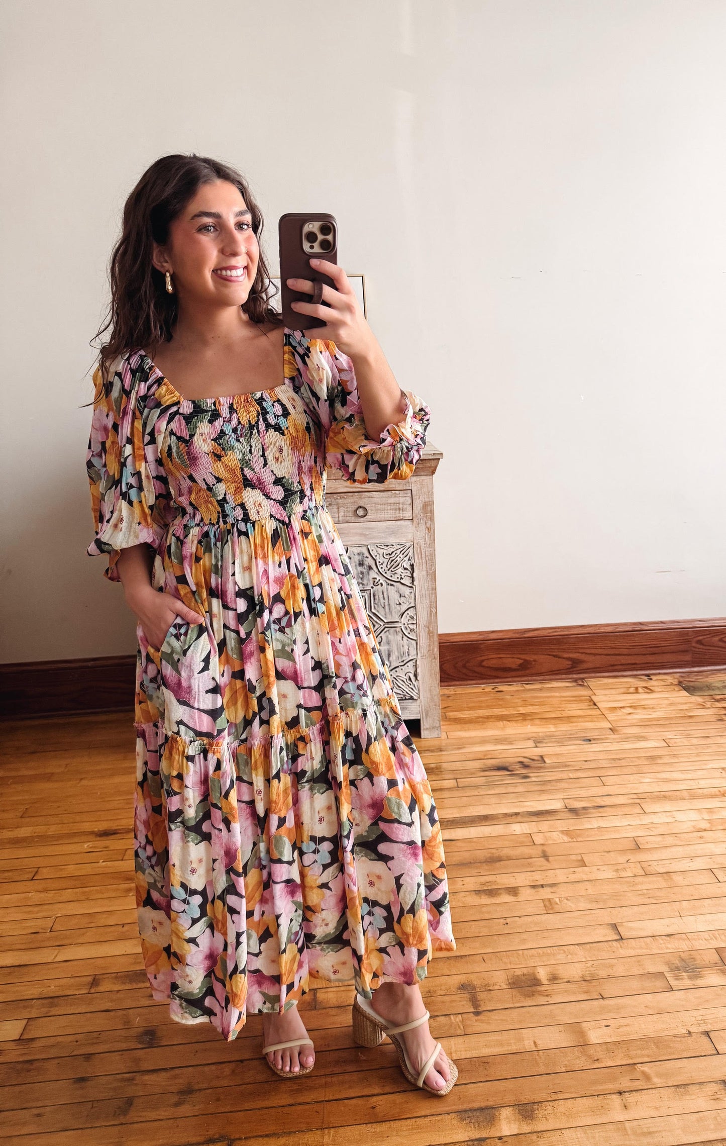 Woman taking a mirror selfie wearing a floral dress in a room with wooden flooring.