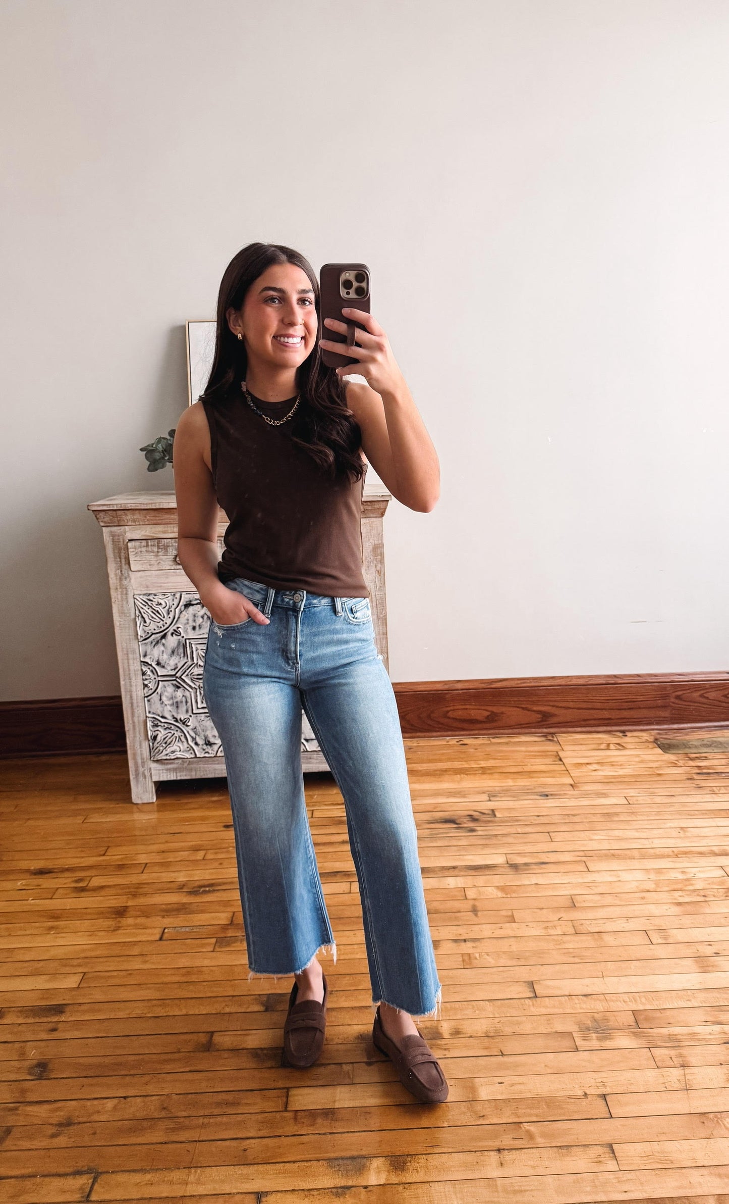 Woman taking a mirror selfie wearing a brown top and light blue jeans in a room with wooden flooring.