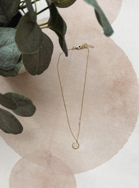 A delicate gold chain necklace with a small open circle pendant, styled on a textured beige surface with soft pink watercolor circles and green eucalyptus leaves.