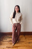 A woman stands in a room with a light gray wall and wooden floor, wearing a cream ribbed knit sweater and dusty rose wide-leg drawstring pants.
