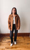 A woman stands in a room with a white wall and wooden floor, wearing a brown corduroy jacket over a white top, black jeans, and white sneakers.