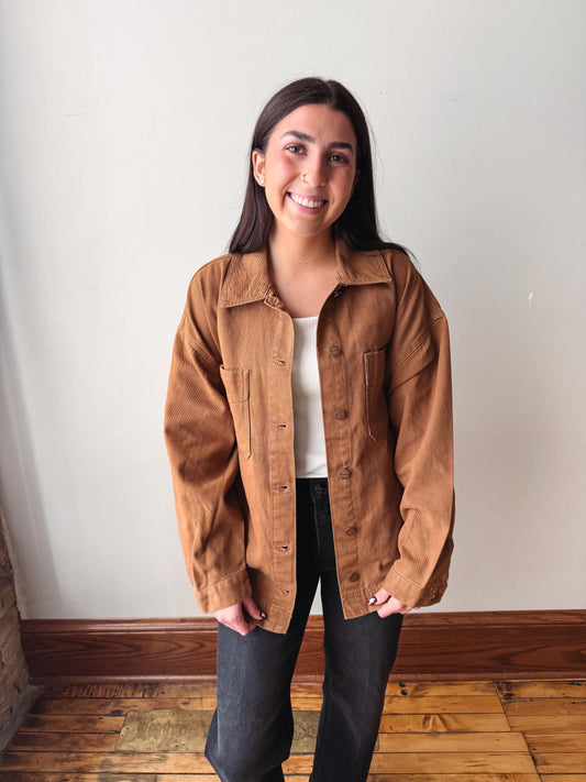 A woman smiling while wearing a brown corduroy button-up jacket over a white top and black pants, standing in front of a plain off-white wall with wooden flooring.