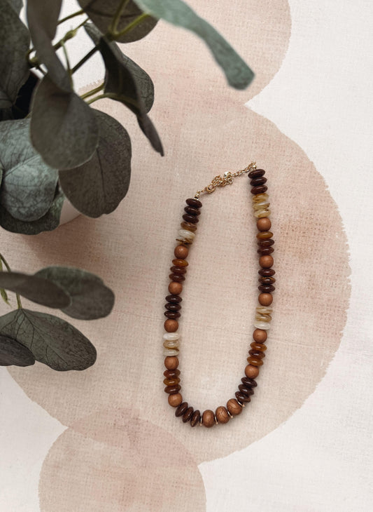 A beaded necklace with dark brown, medium brown, and cream beads, styled on a light pink textured fabric with eucalyptus leaves in the corner.