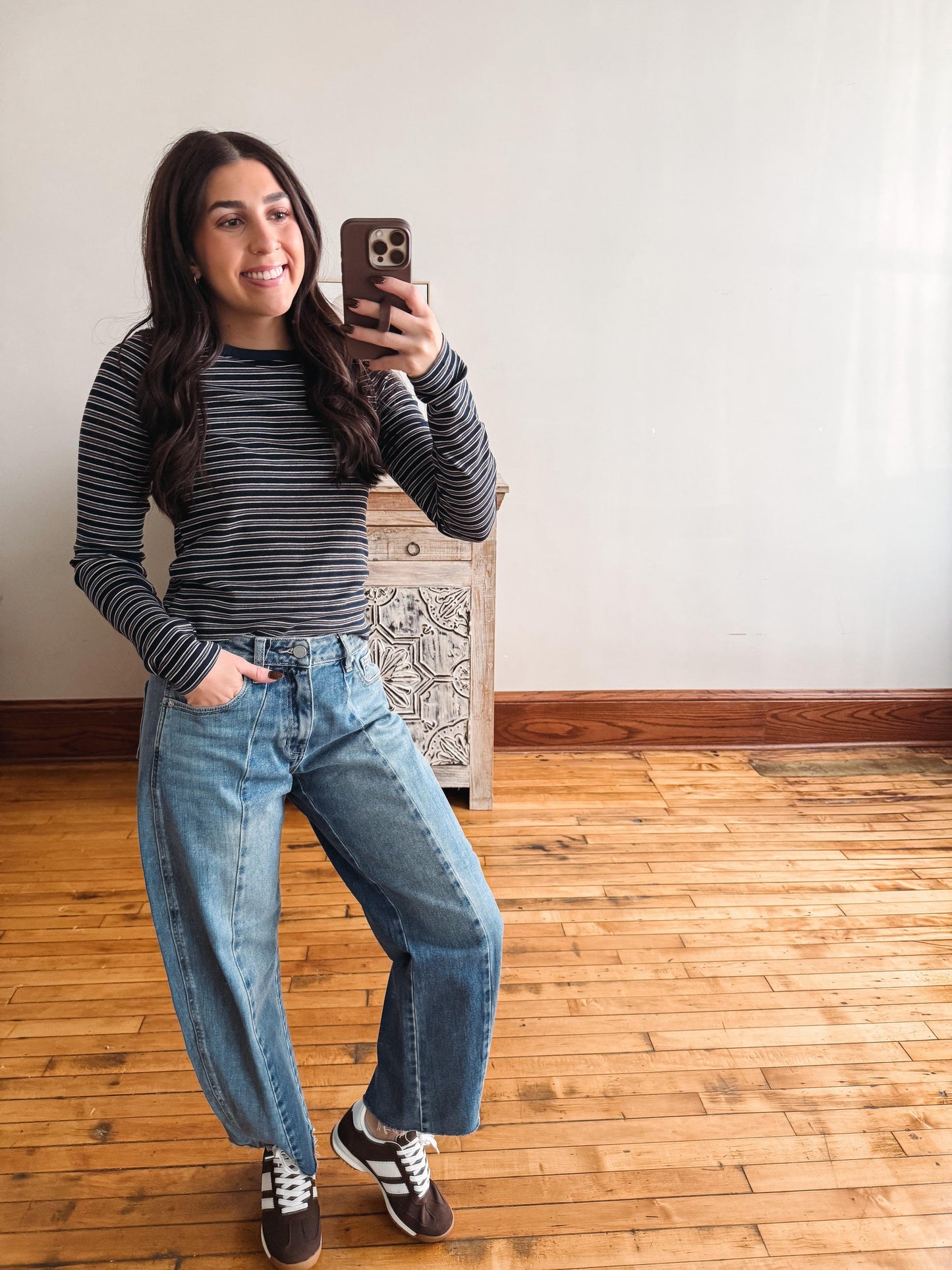 A woman takes a mirror selfie wearing a black and gray striped long-sleeve shirt, wide-leg blue jeans, and brown sneakers with white laces, standing in a room with a light-colored wall and wooden floor.