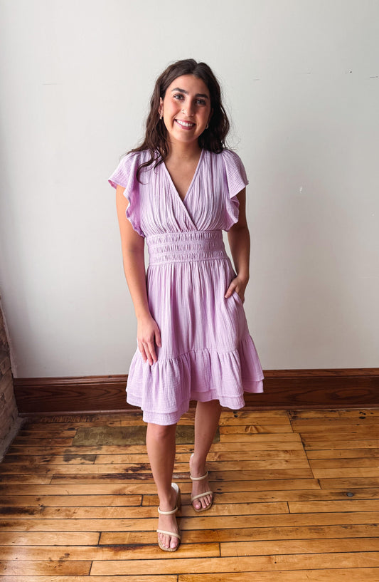 Woman wearing a light purple dress standing on a wooden floor with a white wall background