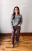 A woman stands in a room with a light gray wall and wooden floor, wearing a gray ribbed knit sweater with thin white horizontal stripes, brown wide-leg pants, and taupe ankle boots.