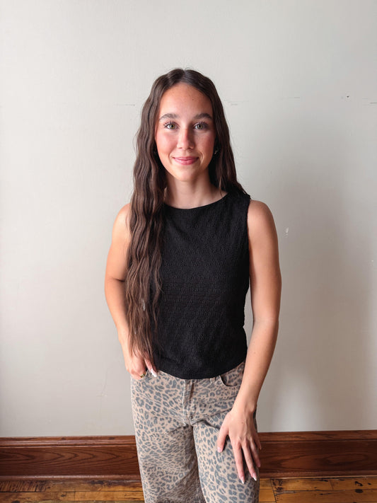 Woman wearing a black sleeveless top and patterned pants against a plain wall.