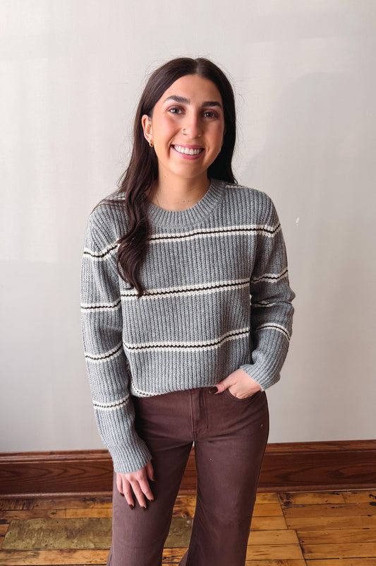 A woman smiling and wearing a gray knit sweater with horizontal cream stripes and brown pants, standing against a plain off-white wall with a wooden baseboard and wood floor.