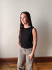 Woman wearing a black sleeveless top and patterned pants standing against a plain wall.