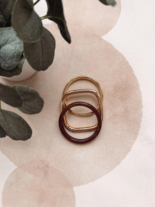 Three small interlocking ring-shaped accessories in gold, rose gold, and dark burgundy, arranged on a pale pink textured surface with decorative green leaves in the corner.