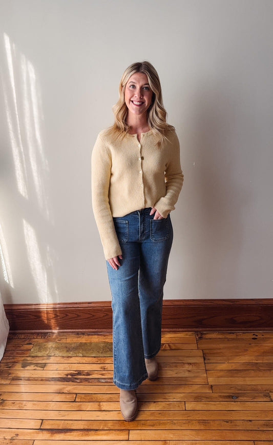 Woman wearing a light yellow cardigan and blue jeans standing in a room with wooden flooring and a white wall.