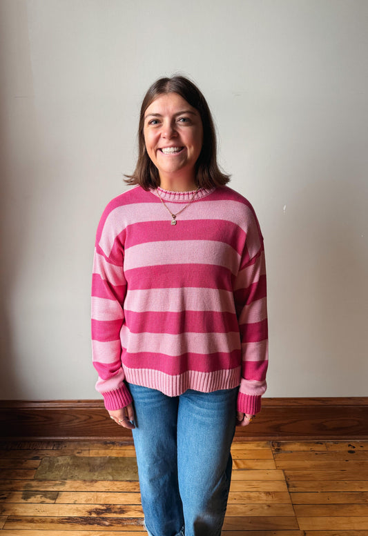 A smiling woman with shoulder-length brown hair stands indoors wearing a pink and light pink horizontally striped knit sweater and blue jeans against a plain off-white wall and wooden floor.