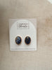 A pair of oval stud earrings with dark blue and white marbled stones in gold bezel settings, displayed on a white arched backing card labeled Urban Threads, set against a beige linen background.