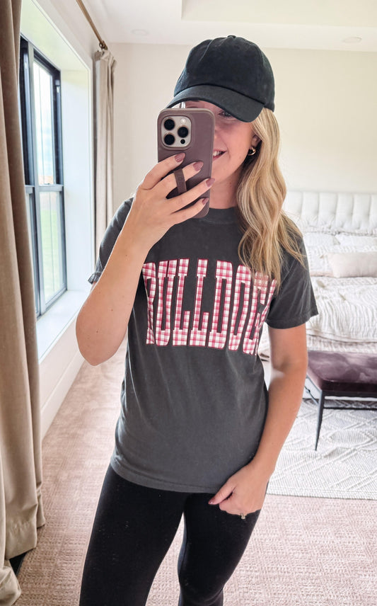 A woman takes a mirror selfie in a bedroom while wearing a dark gray t-shirt with 'BULLDOGS' printed in red and white gingham block letters, paired with a black baseball cap and black leggings.