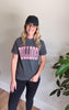 A woman wearing a dark gray graphic t-shirt with 'BULLDOGS' in pink and white block letters, paired with a black baseball cap and black leggings, standing in an indoor setting with a plant in the background.