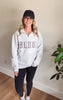 A woman wearing a light gray heathered quarter-zip sweatshirt with 'BULLDOGS' printed across the chest in dark red and gray, paired with a black baseball cap, black leggings, and white sneakers, standing in an indoor setting.