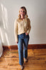 Woman wearing a beige cardigan and blue jeans standing on a wooden floor with a white wall background.