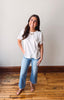 Woman wearing a white t-shirt and blue jeans standing on a wooden floor with a white wall background