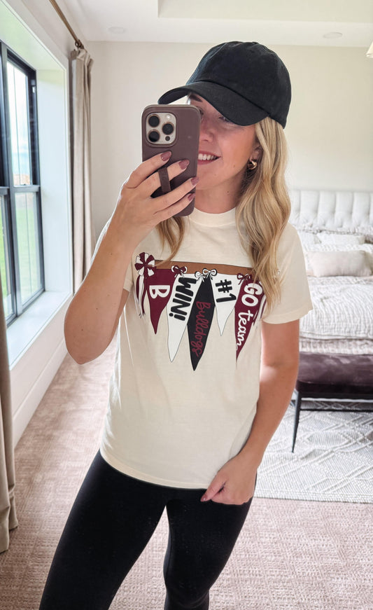 A woman takes a mirror selfie in a bedroom while wearing a cream-colored graphic t-shirt with a maroon and black flag design, a black baseball cap, and black pants.