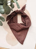 A brown square scarf with small white polka dots, tied in a knot and styled with green eucalyptus leaves on a beige background with abstract circular shapes.