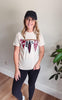 A woman wearing a cream graphic t-shirt with a 'Patriot Sports' bunting design, black leggings, and a black baseball cap, standing in a room with a light beige wall and wood floor.