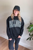 A woman wearing a black crewneck sweatshirt with 'BULLDOGS' printed across the chest in a white pixelated font, paired with a black baseball cap, black leggings, and gray sneakers, standing in an indoor setting with a plant in the background.