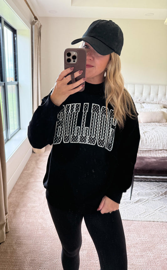 A woman takes a mirror selfie in a bedroom while wearing a black crewneck sweatshirt with 'BULLDOG' printed across the chest in sparkling silver lettering, paired with a black baseball cap and black leggings.