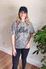 A woman wearing a gray and black acid-wash graphic t-shirt with 'bulldogs' printed in black script, paired with black leggings and a black baseball cap, standing in an indoor setting with a plant in the background.