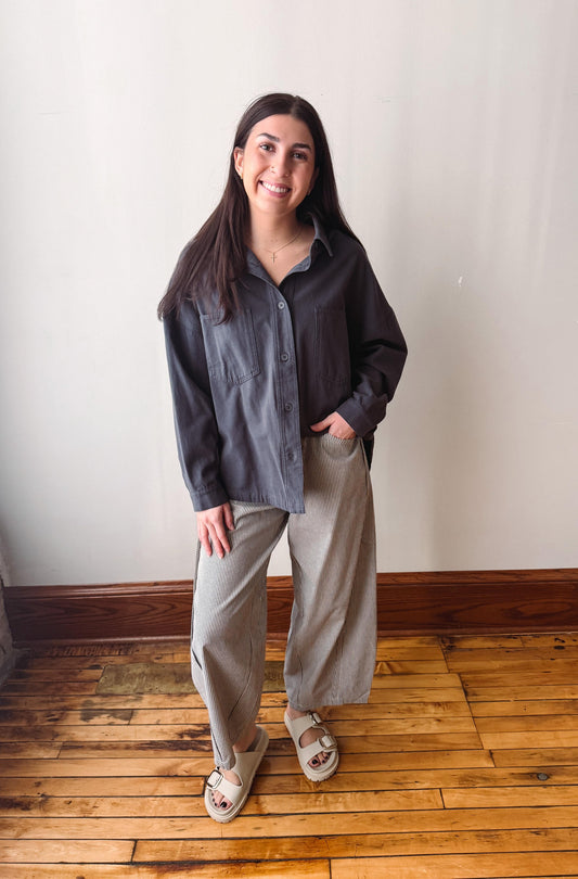 A woman modeling a dark charcoal gray button-down shirt and light beige wide-leg pants, standing on a wooden floor against a plain off-white wall.