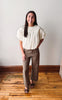 A woman stands in a room with a white wall and wooden floor, wearing a cream puff-sleeve blouse and brown wide-leg pants.