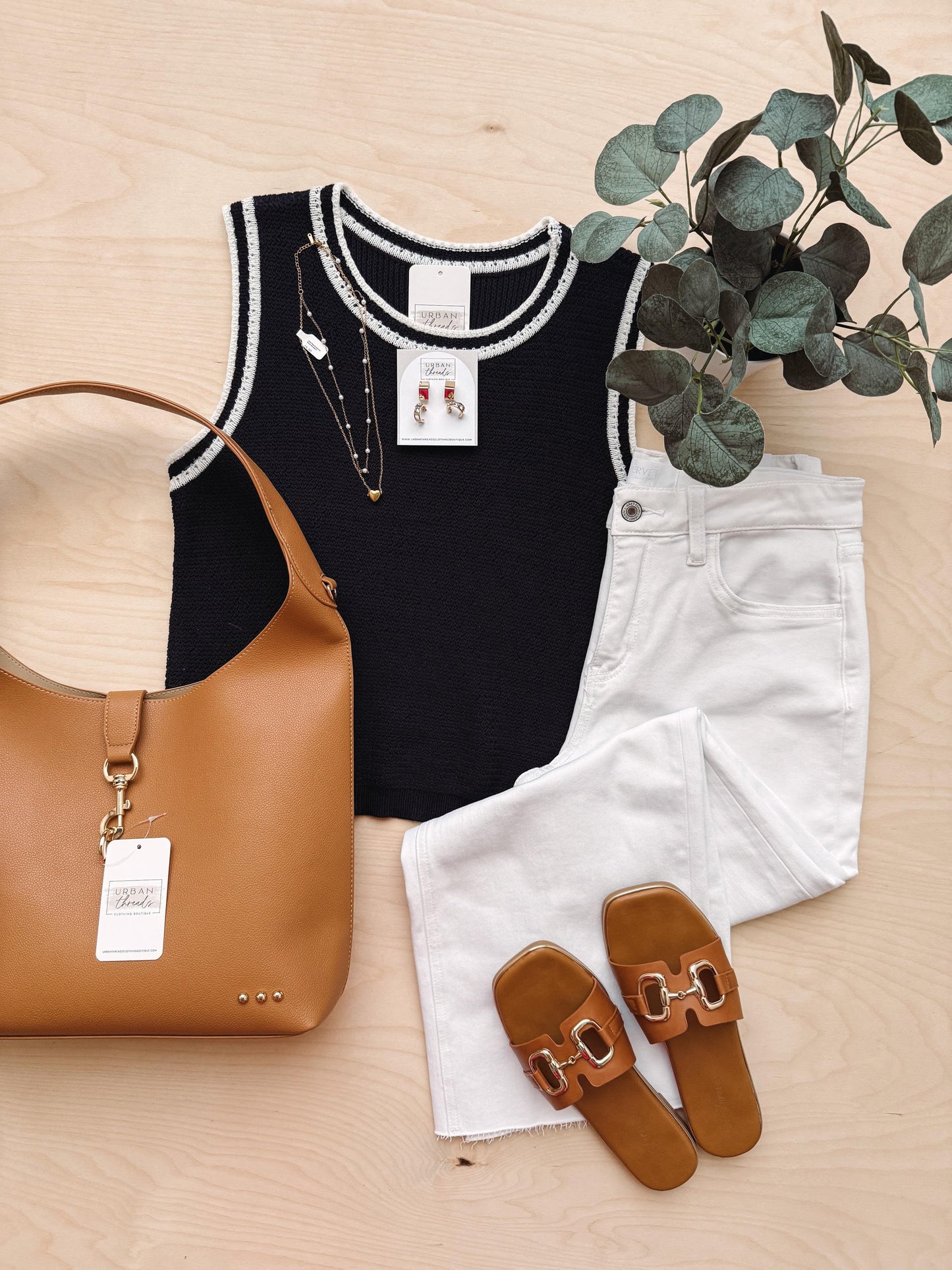 Black sleeveless top with white trim, white shorts, brown sandals, and a tan handbag on a wooden surface with greenery.