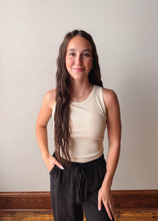 Woman wearing a beige tank top and black pants against a plain wall.