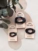 Three black flat cloud hair clips on pink retail cards, styled with green leaves on a light beige surface.