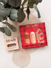 A styled flat-lay of a Kit schü hair perfume discovery set, featuring a red plaid box with two bottles and a separate beige box, arranged on a beige fabric surface with a plant in the background.