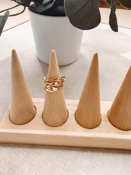 A gold chain-link ring displayed on a light wood cone ring holder, set on a neutral surface with a white planter in the background.